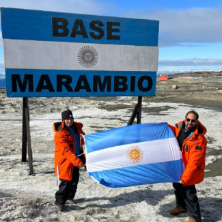 Dos personas desplegando la bandera argentina en la Antártida, delante de un cartel que dice Base Marambio.